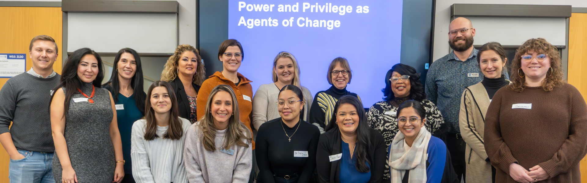 A Group of individuals standing in two rows. Behind them is a screen that says Power and Privilege and Agents of Change